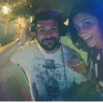 DJ e amica sorridenti vicino alla consolle durante una festa con musica dal vivo e luci colorate.