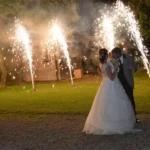 : Sposi che ballano all’aperto di notte tra scintille luminose e fuochi d’artificio durante il matrimonio.