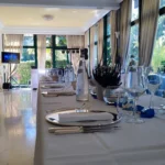 Tavolo elegante con piatti, bicchieri e decorazioni floreali in una sala luminosa preparata per un ricevimento di matrimonio.